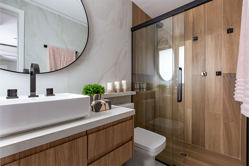 Projeto: Daniela Funari | No banheiro master, materiais naturais criam clima de spa. O espelho redondo iluminado se destaca, e o porcelanato marmorizado off white nas paredes harmoniza com o amadeirado do box