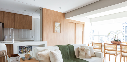 Portas mimetizadas e paleta de cores com os tons de off white e amadeirado claro dão a tônica para reforma de apartamento Projeto: Daniela Funari | Propositalmente, a marcenaria incorporou uma tonalidade que se aproximou ao natural. Na proposta da arquiteta Daniela Funari, essa decisão se complementou com as nuances de off white presentes no piso, sofá, tapete e ilha da cozinha.