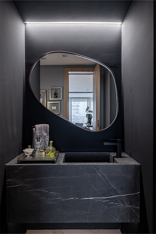 Projeto: Spaço Interior Arquitetura | Escondido atrás de uma porta mimetizada, o lavabo assinado por Ana Rozenblit surpreende com seu design ousado: pórtico preto em drywall, bancada de mármore esculpido e espelho orgânico sob luz indireta.