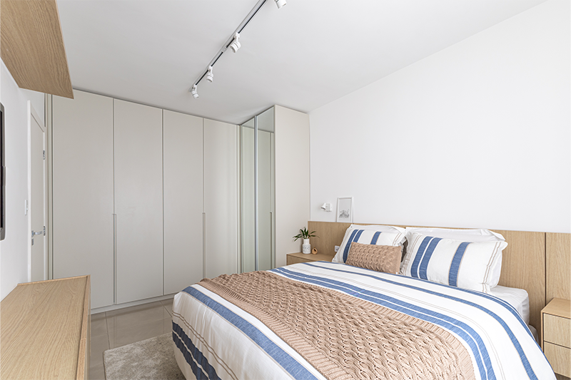 Projeto: Bima Arquitetura | No quarto de casal, marcenaria clara em L e cabeceira de madeira com iluminação embutida unem conforto e funcionalidade