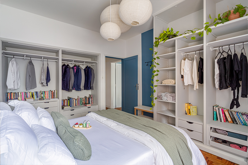 Projeto: Julia Otaga | No quarto do casal, o armário aberto em marcenaria sob medida organiza roupas, livros e objetos pessoais de forma prática e acolhedora. A pintura azul destaca portas e paredes, enquanto o uso de plantas traz frescor e leveza ao ambiente
