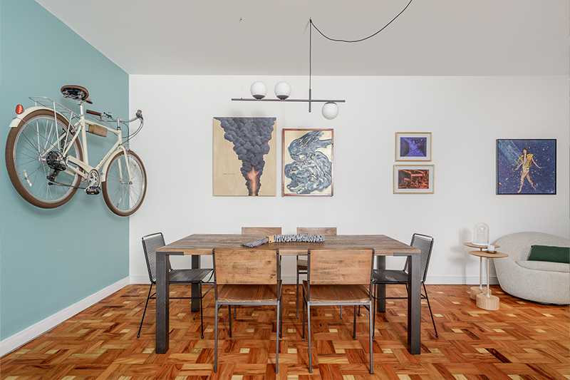 Projeto: Julia Otaga | Na sala de jantar, a marcenaria em madeira dialoga com o piso em tacos, enquanto a parede em tom de azul e a bicicleta suspensa trazem personalidade. Obras de arte e design contemporâneo completam a atmosfera criativa e acolhedora do ambiente.
