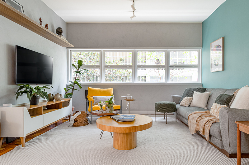 Projeto: Julia Otaga | Na sala de estar, tons neutros ganham vida com o contraste do azul na parede e a poltrona amarela Emmet, que se torna ponto de destaque. A marcenaria leve, as plantas e a iluminação natural criam um espaço acolhedor e cheio de personalidade.