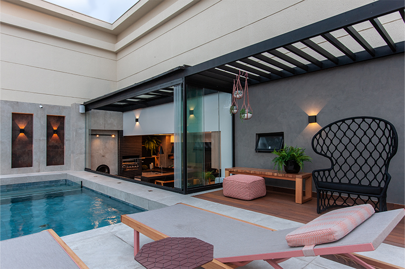 Projeto: Daiane Antinolfi | A piscina integrada ao terraço conta com divisória em vidro e pergolado preto, criando fluidez entre os ambientes de lazer.