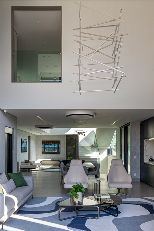 Projeto: Érica Salguero | O pé-direito duplo valoriza a integração do estar e jantar, com pendente escultural e tapete em azul que conecta o interior ao clima praiano. Já a piscina, integrada à área social, reforça a atmosfera de lazer e bem-estar, tornando-se ponto de encontro da casa.