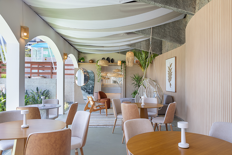 Projeto: Rodrigo Coelho | Mostra Morar Mais 2025 | Um grande painel ripado em forma de onda emoldura a parede de pedras, enquanto o teto em tecido completa a composição com leveza.