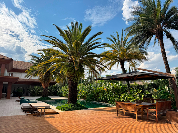 Ao fundo da piscina há um jardim tropical denso que foi executado especialmente para uma moradora apaixonada por plantas. O verde das espécies contribui para uma paisagem mais natural em meio ao piso de travertino e o deck de madeira.