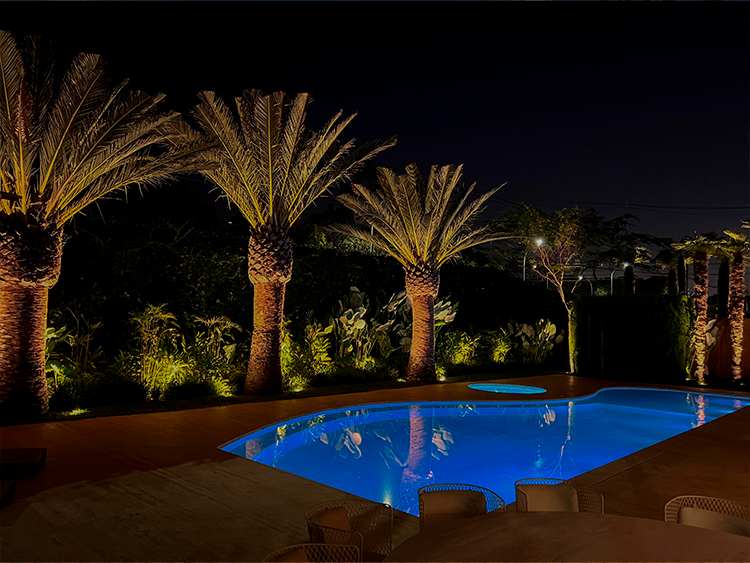 Mais um exemplo de jardim com vegetação tropical e palmeiras do tipo Canariense. A imagem noturna do projeto traz a visão da piscina iluminada, cenário ideal para festas e encontros especiais.