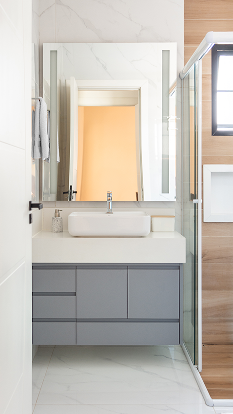 No banheiro, a marcenaria cinza se harmoniza com revestimentos claros e porcelanato amadeirado no box, enquanto a iluminação embutida valoriza os materiais e reforça a atmosfera atual da casa 
